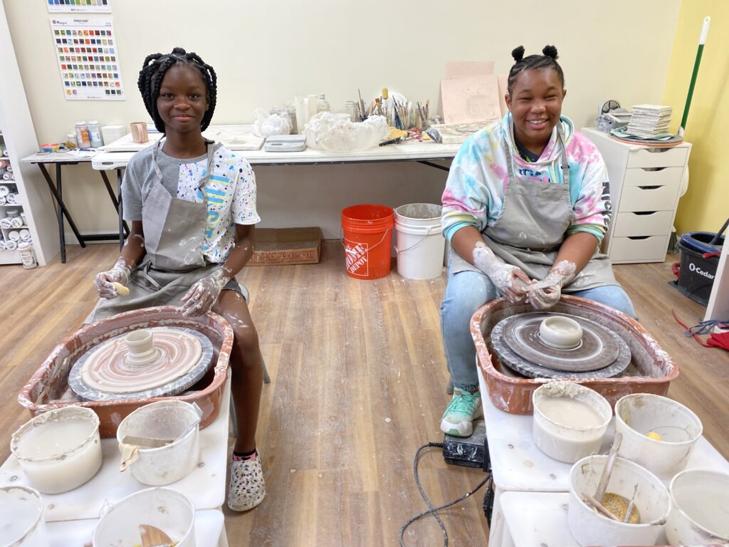two girls sit at the pottery wheel with pieces on the wheel