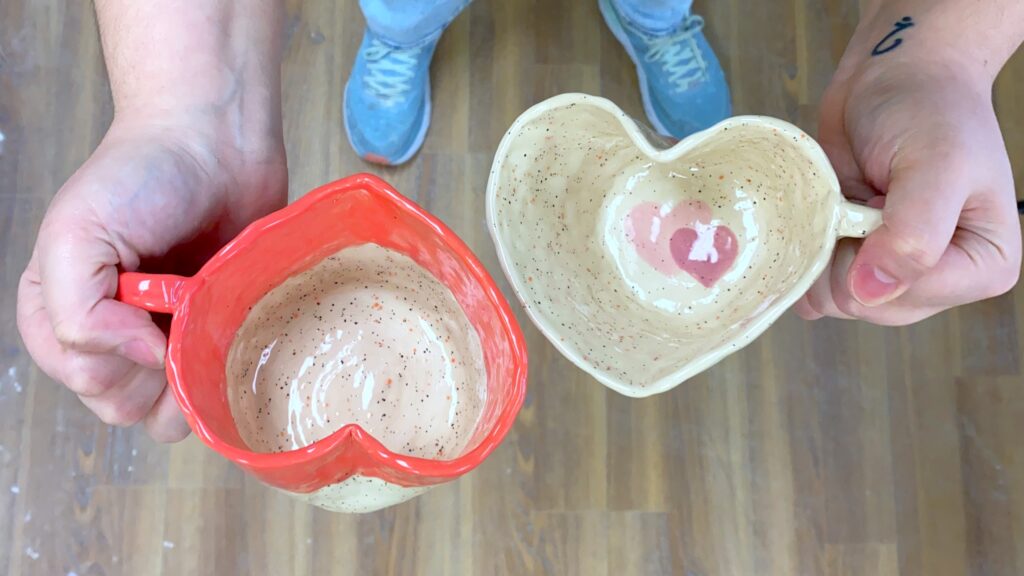 two mugs in the shape of hearts held in two hands. they are painted differently, one with speckles and the other with an orange rim.