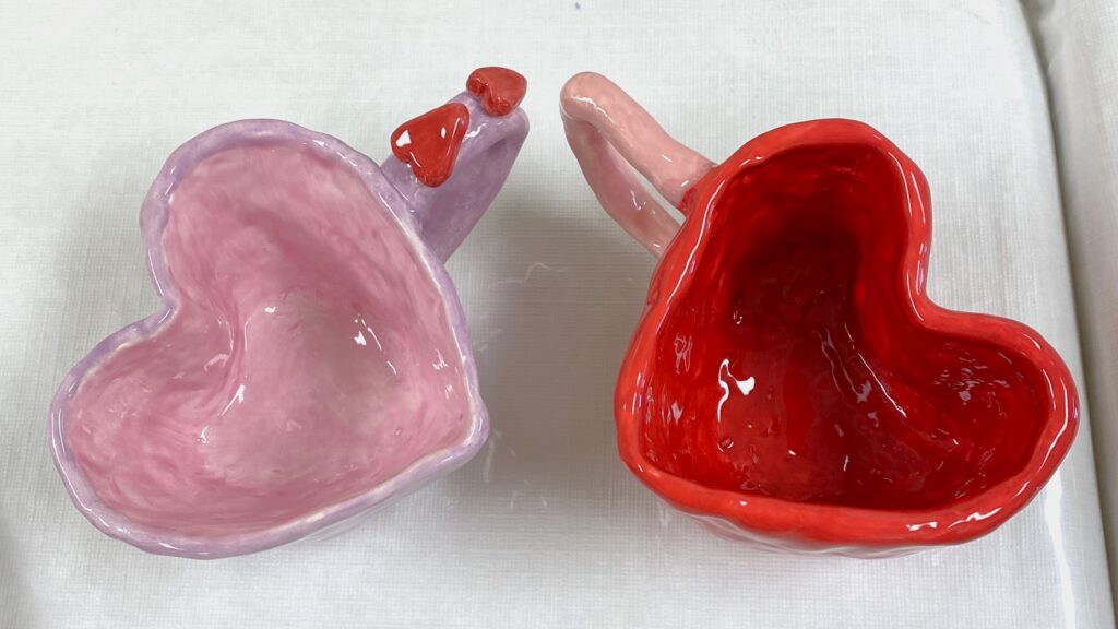 two mugs in the shape of hearts. one mug has hearts on the handle. they are painted pink and red.