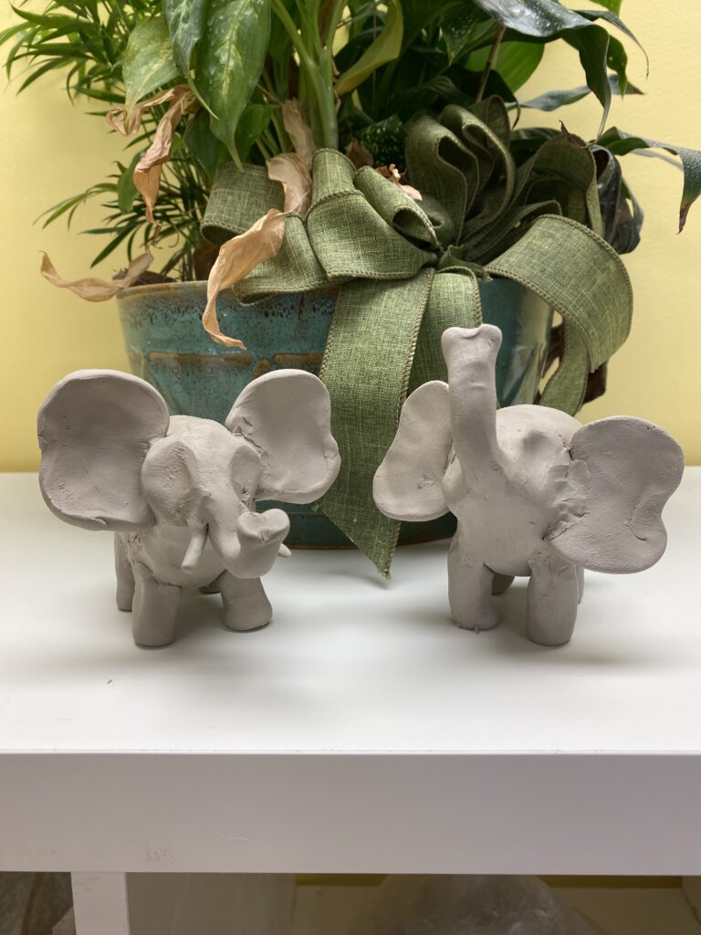 two unfired clay elephants in front of a pot with a plant in it