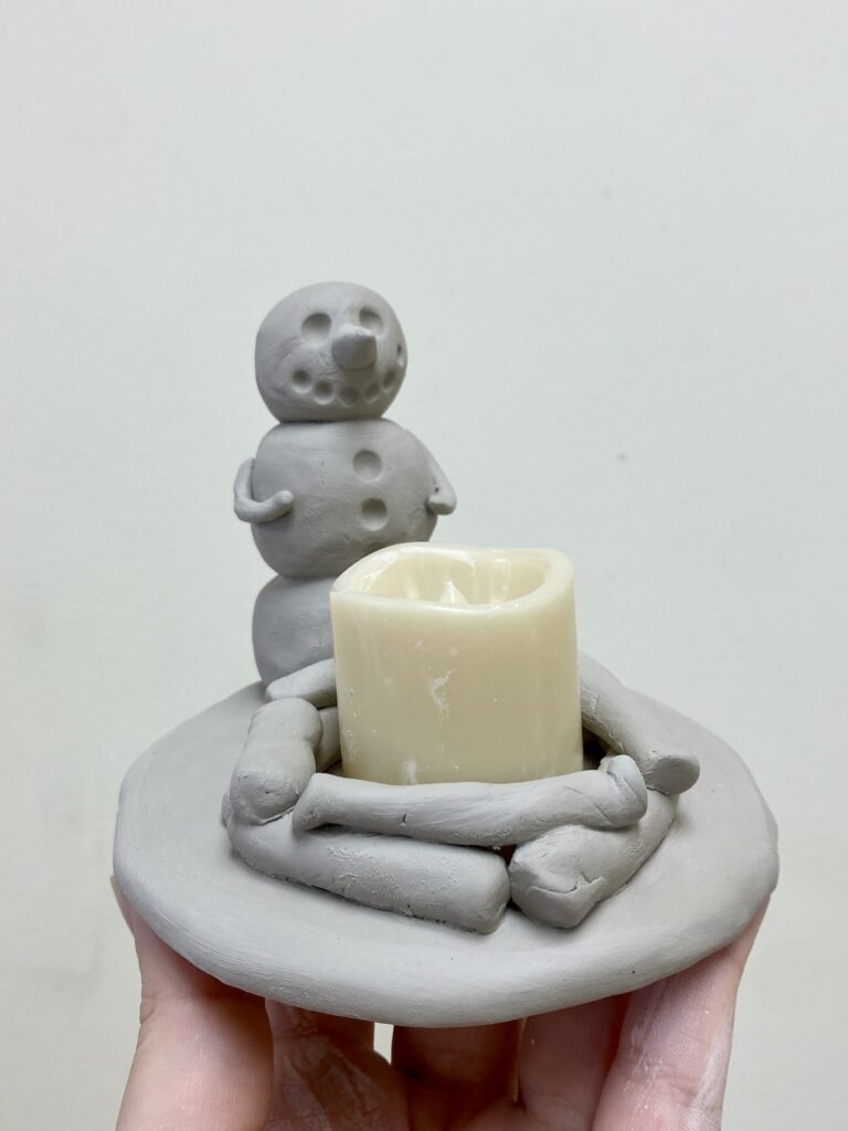 clay snowman sitting in front of a fire pit where the fire pit would fit a tealight candle holder, and there is a tealight candle resting there