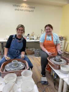 two women smiling sitting at the pottery wheel with clay formed into a vase on the wheel