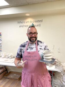 man holding a freshly thrown vase from the pottery wheel, smiling and pointing to the vase sitting on a wooden plate 