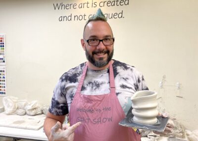 man holding a freshly thrown vase from the pottery wheel, smiling and pointing to the vase sitting on a wooden plate
